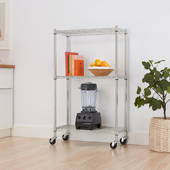A chrome three-tier wire rolling cart sits in a light-filled kitchen space next to a potted houseplant and white cabinetry. The shelves are neatly arranged with colorful cookbooks, a bowl of fresh citrus fruits, and a black countertop blender stored on the bottom tier.
