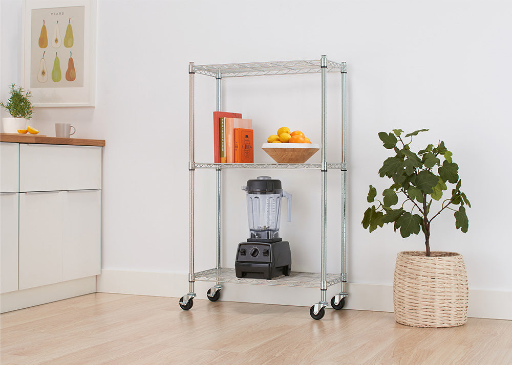 A chrome three-tier wire rolling cart sits in a light-filled kitchen space next to a potted houseplant and white cabinetry. The shelves are neatly arranged with colorful cookbooks, a bowl of fresh citrus fruits, and a black countertop blender stored on the bottom tier.