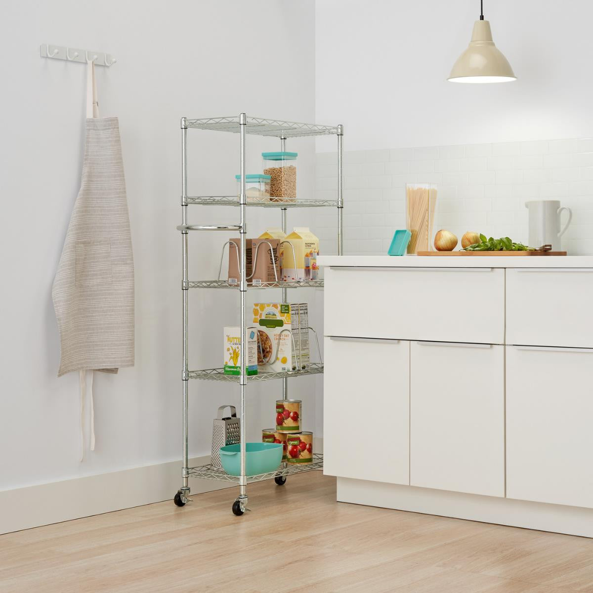 A tall, chrome wire shelving unit with five tiers stands in a kitchen, filled with various pantry items like cereal boxes, canned goods, and food containers. To the left, a beige and white striped apron hangs on a white wall with a row of hooks.