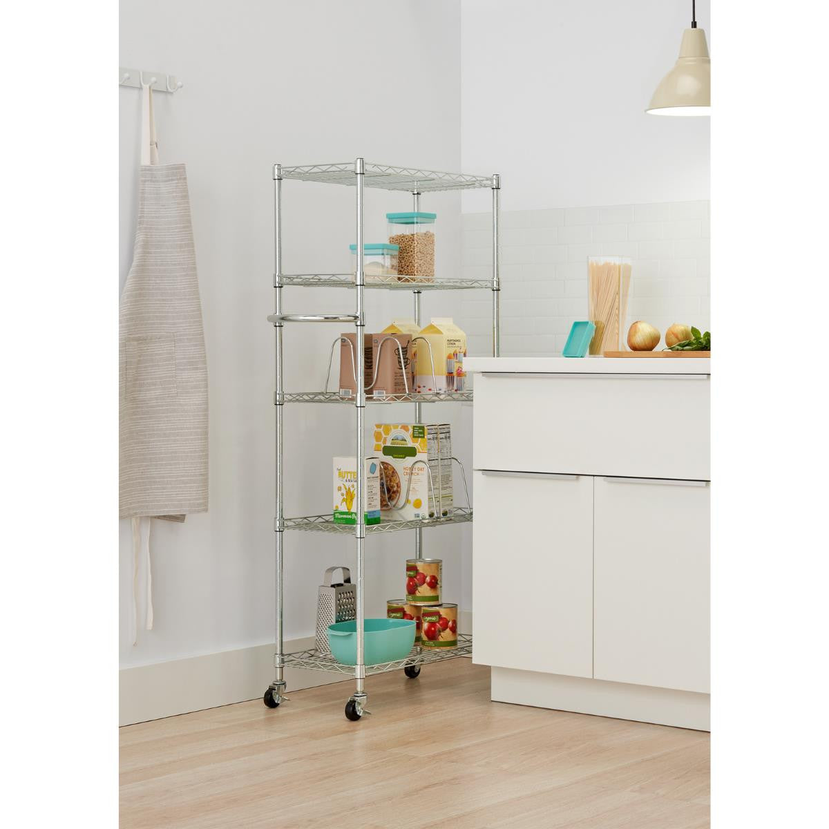 A tall, narrow, silver metal wire shelf unit with five tiers on wheels sits in a kitchen. The shelves hold various pantry items, including jars, boxes of food, cans, and a blue bowl.