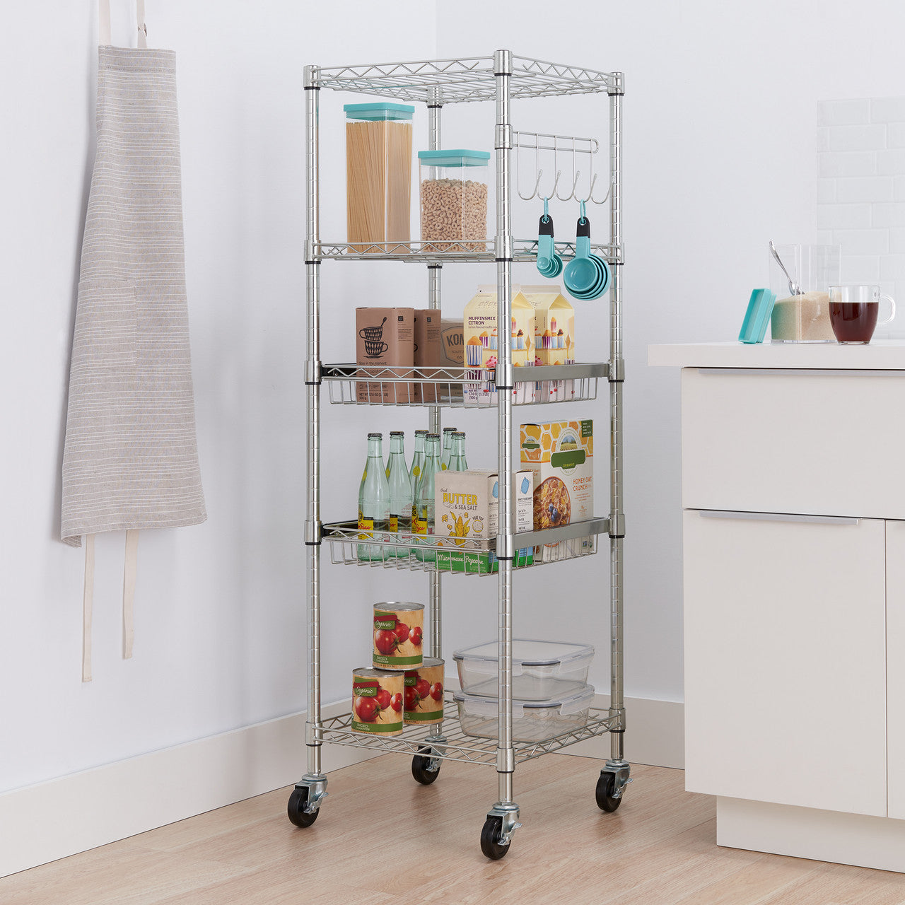 A tall, five-tier chrome wire rack stands in a kitchen corner, filled with various pantry items. The rack has wheels for mobility and includes both wire shelves and two black wire baskets.