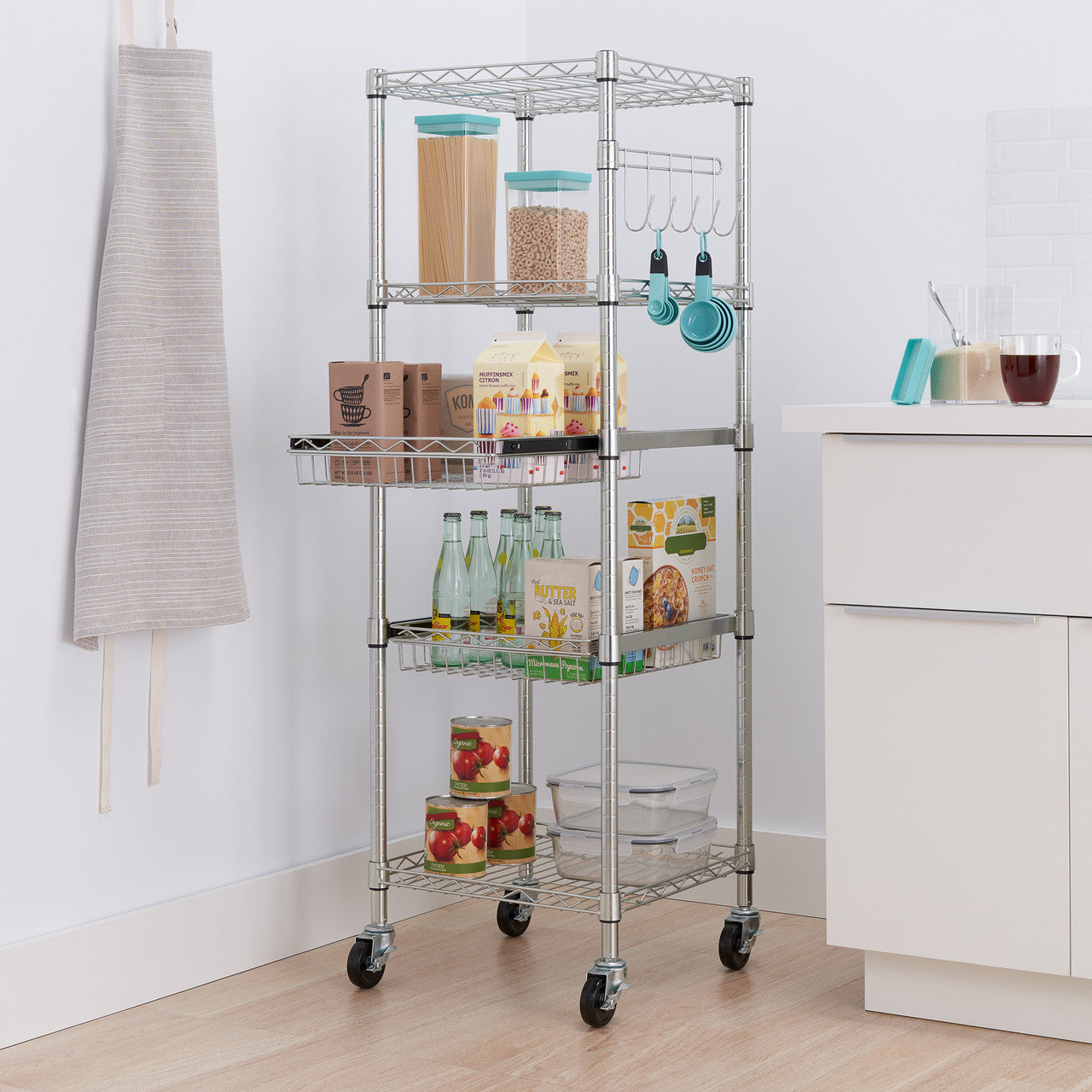 A tall, chrome wire shelving unit with five tiers stands in a kitchen setting. The rack holds various pantry items, including jars of grains, boxes of food, bottles, cans, and clear storage containers, demonstrating an organized storage solution.