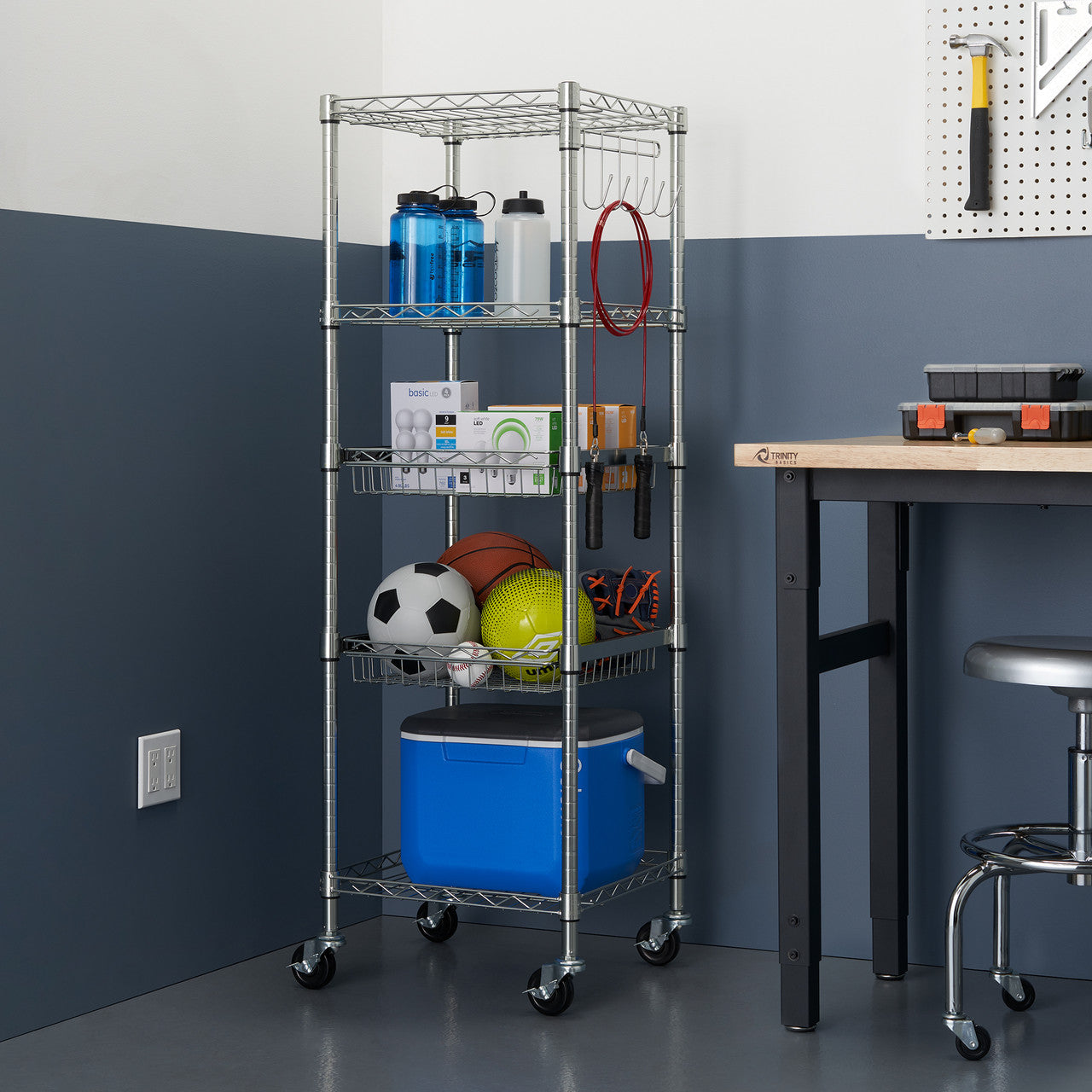 A five-tier Trinity EcoStorage chrome rack stands against a two-toned wall. The rack holds various items, including water bottles, sports equipment, and a blue cooler.