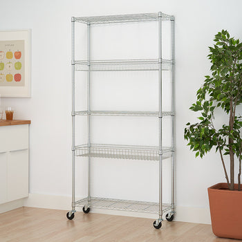 A silver, 5-tier wire shelving rack on wheels stands against a white wall, positioned between a white cabinet and a potted plant with green leaves. The rack has four shelves with a wire basket design and one flat wire shelf at the bottom.