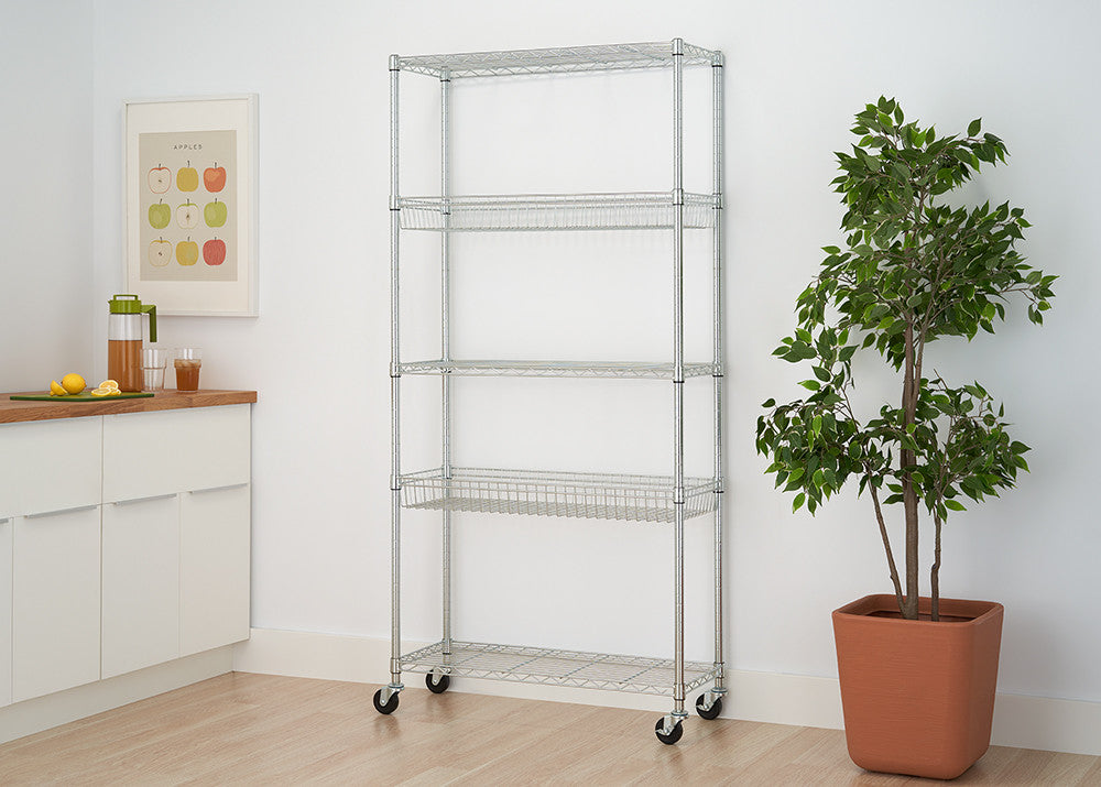 A silver, 5-tier wire shelving rack on wheels stands against a white wall, positioned between a white cabinet and a potted plant with green leaves. The rack has four shelves with a wire basket design and one flat wire shelf at the bottom.