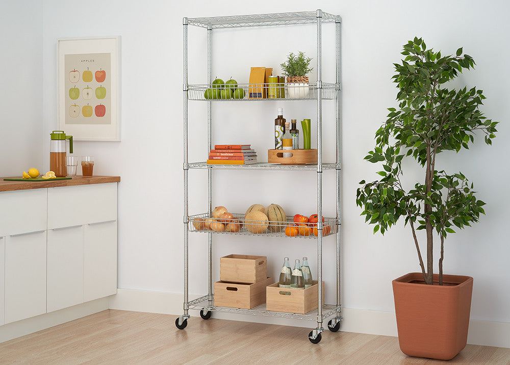 A silver, 5-tier wire shelving unit with wheels is positioned against a white wall. The shelves hold various items including fruits, books, and decorative objects, and the unit is flanked by a potted plant and a kitchen counter.