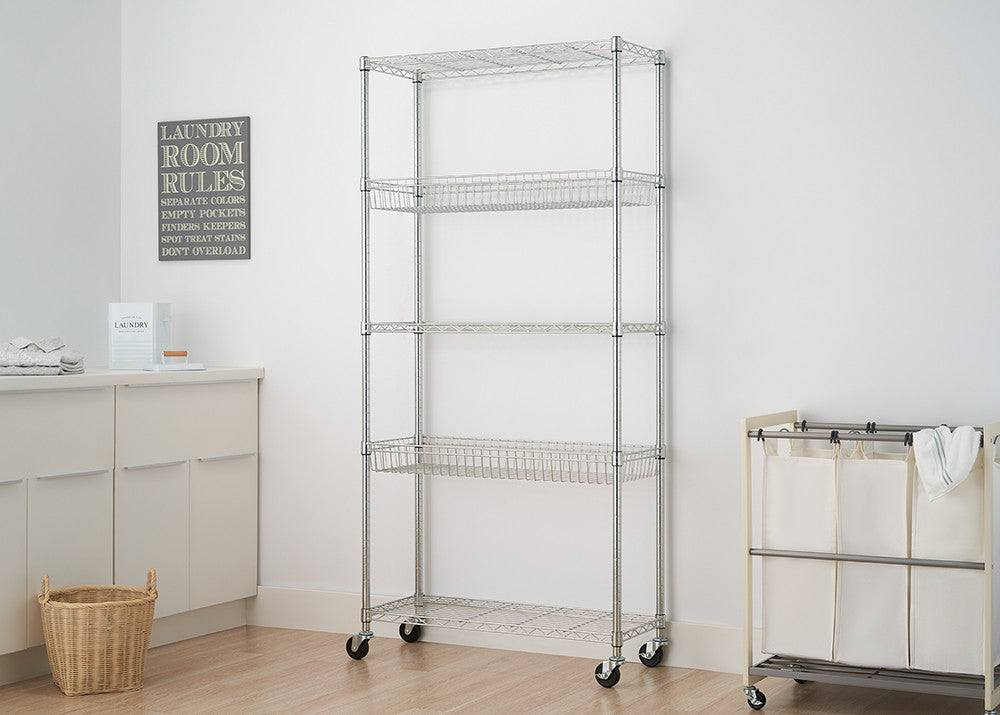 A 5-tier wire shelving rack with baskets and wheels stands against a white wall in a laundry room. A laundry basket sits on the floor to the left, and a rolling laundry sorter is on the right.