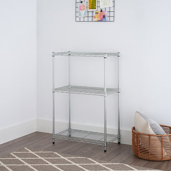 A low-angle shot of a three-tier wire shelving rack with a chrome finish, positioned in a room with a light gray wall and a patterned rug. A woven basket with a pillow is placed to the right of the shelving unit.