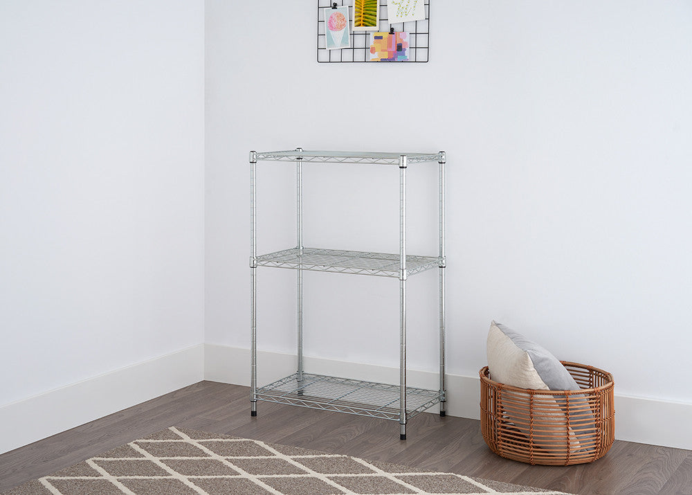 A low-angle shot of a three-tier wire shelving rack with a chrome finish, positioned in a room with a light gray wall and a patterned rug. A woven basket with a pillow is placed to the right of the shelving unit.