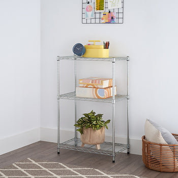A silver, three-tiered wire shelving unit stands in the corner of a room with white walls, holding boxes, a plant, and a yellow container. A woven basket with pillows sits beside the shelving unit, and a patterned rug is visible on the floor.