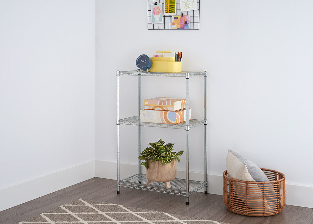 A silver, three-tiered wire shelving unit stands in the corner of a room with white walls, holding boxes, a plant, and a yellow container. A woven basket with pillows sits beside the shelving unit, and a patterned rug is visible on the floor.