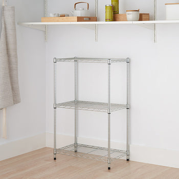 A silver, three-tier wire shelving unit stands on a light wood floor against a white wall. The unit has a simple design with three shelves and four legs.