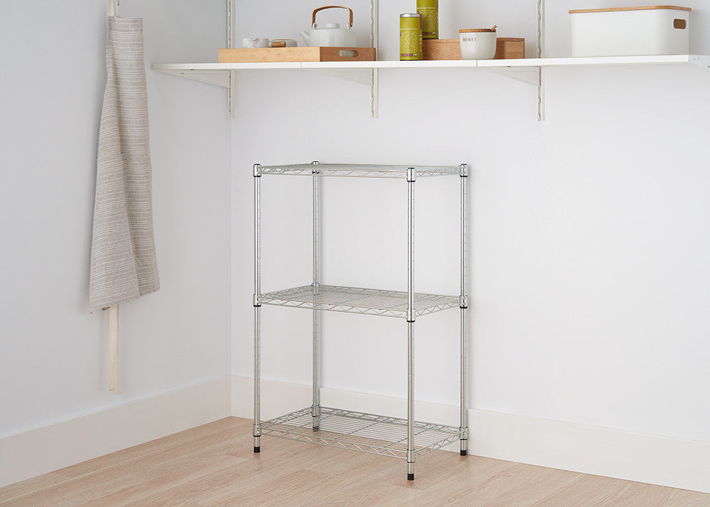 A silver, three-tier wire shelving unit stands on a light wood floor against a white wall. The unit has a simple design with three shelves and four legs.