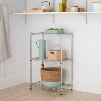 A small, three-tiered metal wire shelving unit stands against a white wall in a room with light wood flooring. The shelves hold a basket, a wooden box with bottles, a pitcher, and a green container with fruit.