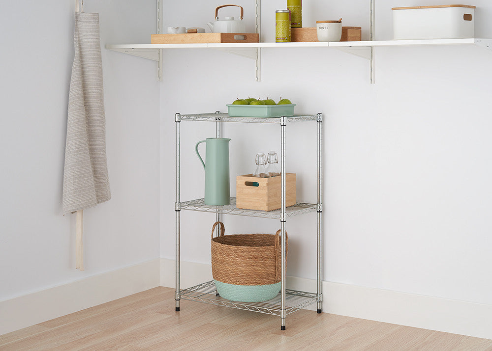A small, three-tiered metal wire shelving unit stands against a white wall in a room with light wood flooring. The shelves hold a basket, a wooden box with bottles, a pitcher, and a green container with fruit.