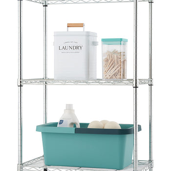 A silver wire shelving unit with three tiers is shown against a white background.  The shelves hold a white laundry bin, a clear container of clothespins, a bottle of detergent, and two round white objects.