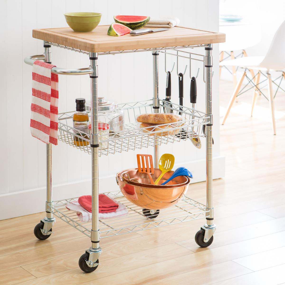 A chrome kitchen cart with a light brown cutting board top is shown in a kitchen setting. The cart holds various kitchen items, including bowls, food, utensils, and towels, and features multiple shelves and hooks.