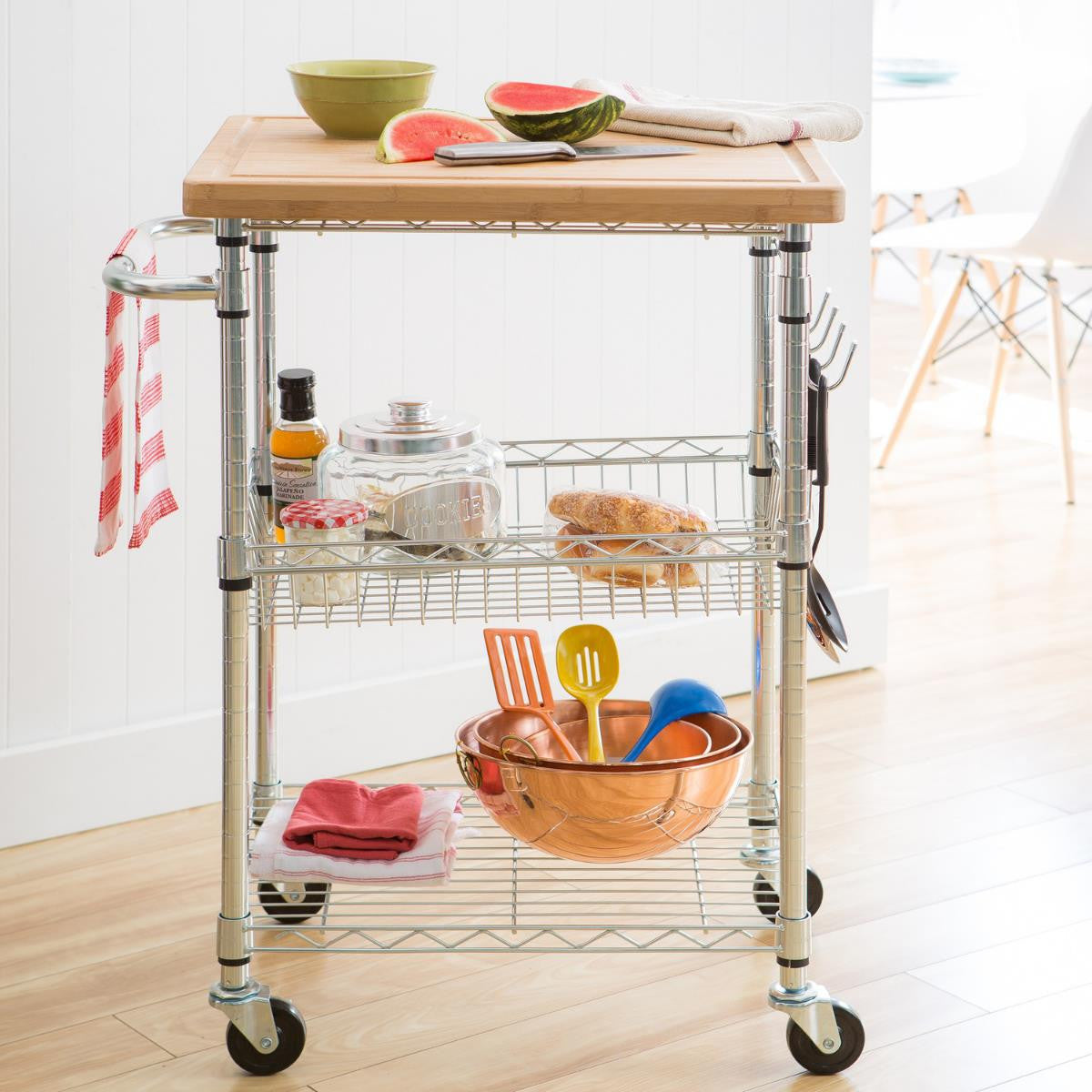 A chrome kitchen cart with a bamboo cutting board on top holds various kitchen items. The cart has three shelves and wheels for mobility.