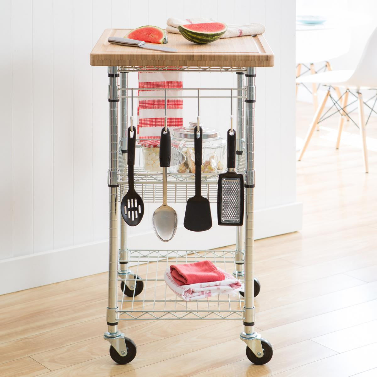A chrome metal kitchen cart with four black wheels stands on a light wooden floor against a white wall. The cart has a light brown wooden cutting board on top with slices of watermelon and a knife, along with two wire shelves holding jars and folded towels, and hooks holding kitchen utensils.