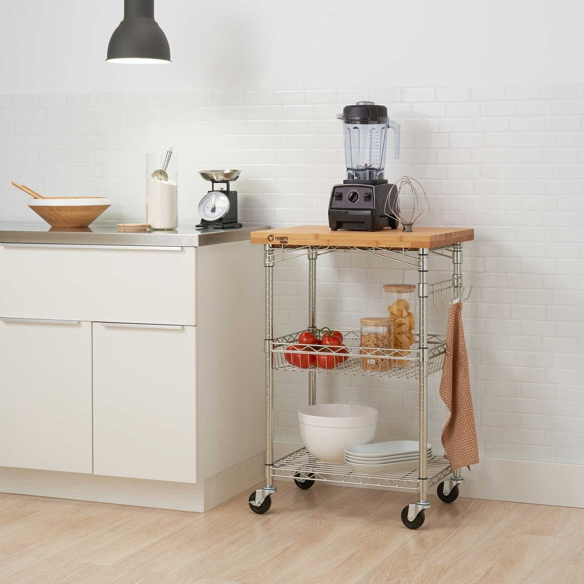 A compact rolling kitchen cart with a wooden top holds a blender and whisk, with tomatoes, jars, bowls, and plates stored on its wire shelves. The cart stands beside white kitchen cabinets against a bright white tiled wall.