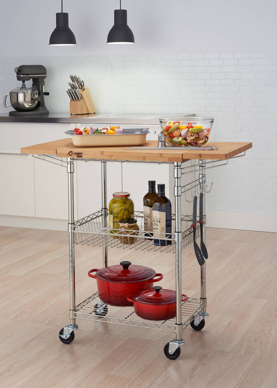 A stainless steel kitchen cart with a light brown bamboo top sits in a kitchen. The cart has two wire shelves holding red pots, jars, and bottles, and utensils hanging from the side.