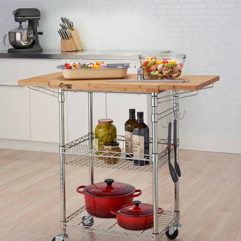 A stainless steel kitchen cart with a light brown bamboo top sits in a kitchen. The cart has two wire shelves holding red pots, jars, and bottles, and utensils hanging from the side.