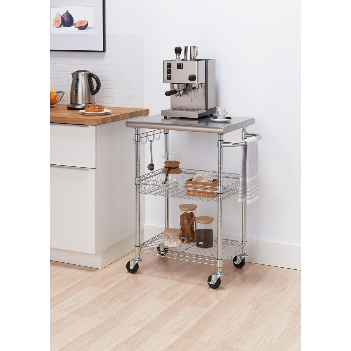 A stainless steel kitchen cart with three tiers stands on a light wood floor against a white wall. The top tier holds an espresso machine and a white cup, while the lower tiers contain jars and containers; a towel hangs from the cart's handle.