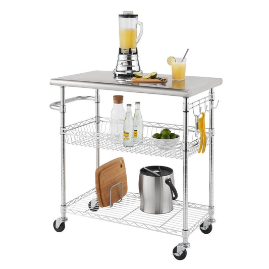 A stainless steel kitchen cart with three shelves on wheels is shown against a white background. The top shelf holds a blender, a cutting board with limes, and a glass of a light-colored drink. The middle shelf contains two wire baskets holding bottles and a bowl of limes, while the bottom shelf has a cutting board in a holder and a metal ice bucket. Utensils hang from hooks on the side of the cart.