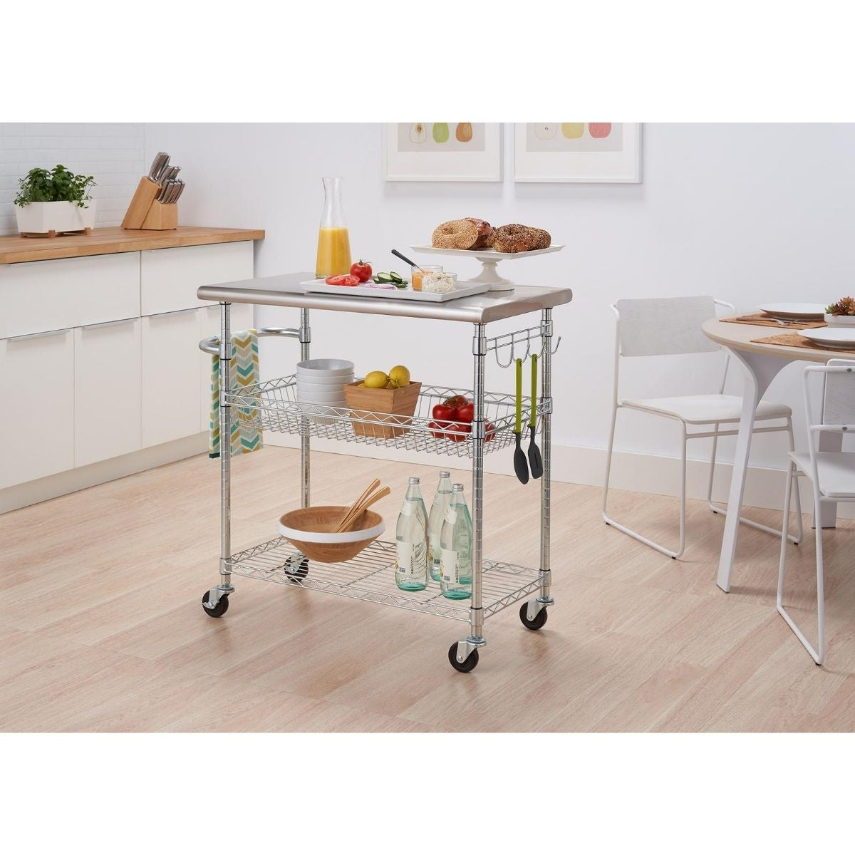 A stainless steel kitchen cart with two wire shelves and a towel rack, holding various kitchen items. The cart is on wheels and positioned in a kitchen setting.