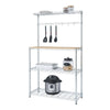 A chrome-colored metal baker's rack with four wire shelves and a light brown wooden countertop is shown against a white background. Kitchen items such as utensils hanging from hooks, jars, and a pressure cooker are arranged on the shelves and countertop.