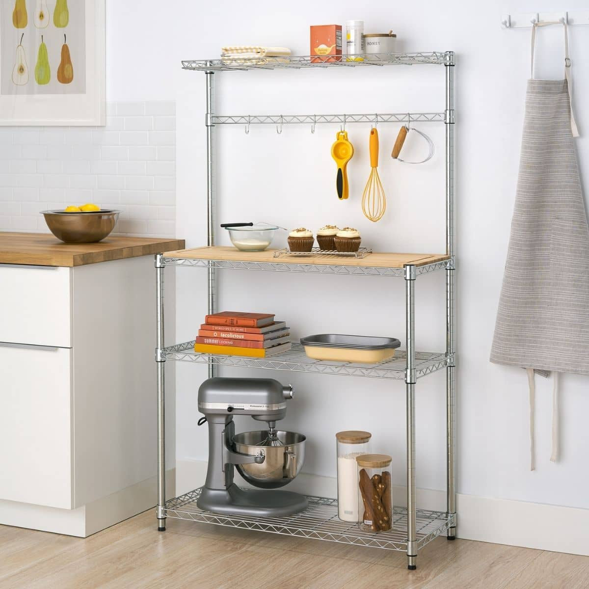 A chrome metal baker's rack stands against a white tiled wall, holding various kitchen items. The rack features wire shelves, a light wood countertop, and hanging hooks with utensils.