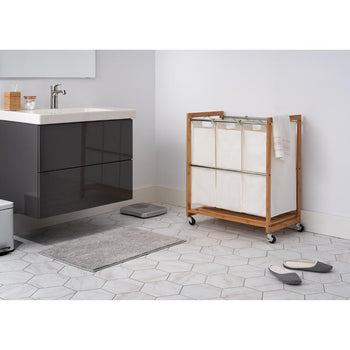 An EcoStorage 3-Bag Bamboo Laundry Cart with white canvas bags sits on a hexagonal tiled floor in a bathroom. A dark grey floating vanity with a white sink and a silver faucet is on the left, and a grey bath mat lies in the center.