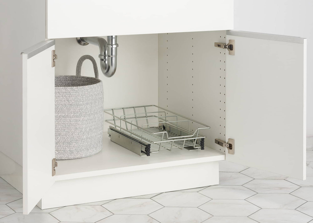 A close-up shows the interior of a white under-sink kitchen cabinet. A chrome wire slide-out drawer on metal tracks is partially extended, sitting next to a textured gray round storage basket with handles.