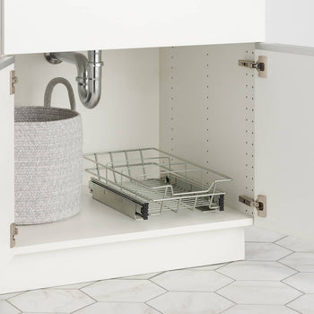 A close-up shows the interior of a white under-sink kitchen cabinet. A chrome wire slide-out drawer on metal tracks is partially extended, sitting next to a textured gray round storage basket with handles.
