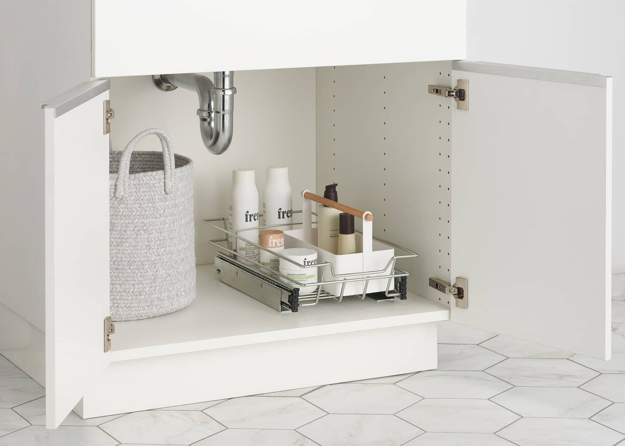 A close-up shows the inside of a white bathroom cabinet under a sink. A light gray oval woven basket with two rope handles sits on the left, while a chrome pull-out organizer with various white bottles and containers rests on the right.