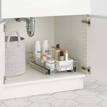 A close-up shows the inside of a white bathroom cabinet under a sink. A light gray oval woven basket with two rope handles sits on the left, while a chrome pull-out organizer with various white bottles and containers rests on the right.