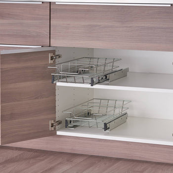 A close-up shows two chrome wire sliding drawers installed within a light brown and white cabinet. Each drawer is empty and extended slightly, revealing the smooth sliding mechanism attached to the cabinet walls.