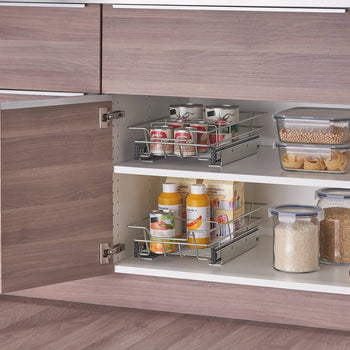 A partially open cabinet reveals two chrome wire sliding drawers installed on white shelves. The drawers contain various pantry items, including canned goods, bottles, and a bag of pasta, illustrating an organized storage solution.
