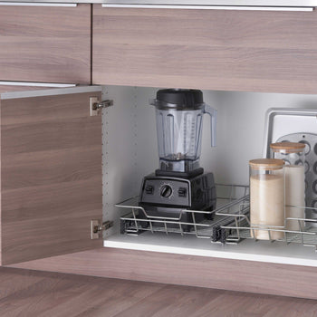 A pull-out chrome wire organizer drawer is installed inside a lower wooden kitchen cabinet with two doors open wide. The sliding rack efficiently stores a black blender, glass storage canisters, a baking sheet, and a muffin pan.