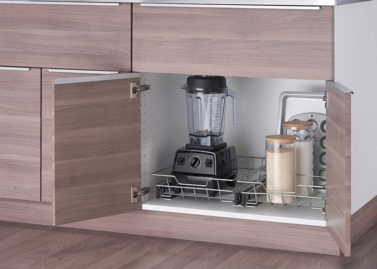 A pull-out chrome wire organizer drawer is installed inside a lower wooden kitchen cabinet with two doors open wide. The sliding rack efficiently stores a black blender, glass storage canisters, a baking sheet, and a muffin pan.