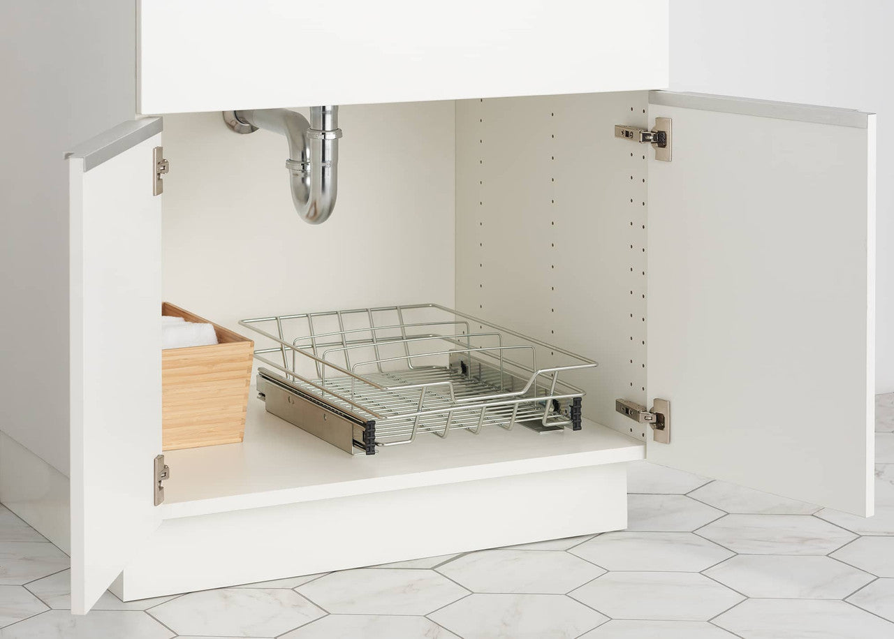 A close-up shows the interior of a white under-sink cabinet with the doors open. Inside, a chrome sliding wire drawer with a handle is partially extended, and a light-colored wooden box filled with white cloths sits to its left.