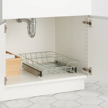 A close-up shows the interior of a white under-sink cabinet with the doors open. Inside, a chrome sliding wire drawer with a handle is partially extended, and a light-colored wooden box filled with white cloths sits to its left.