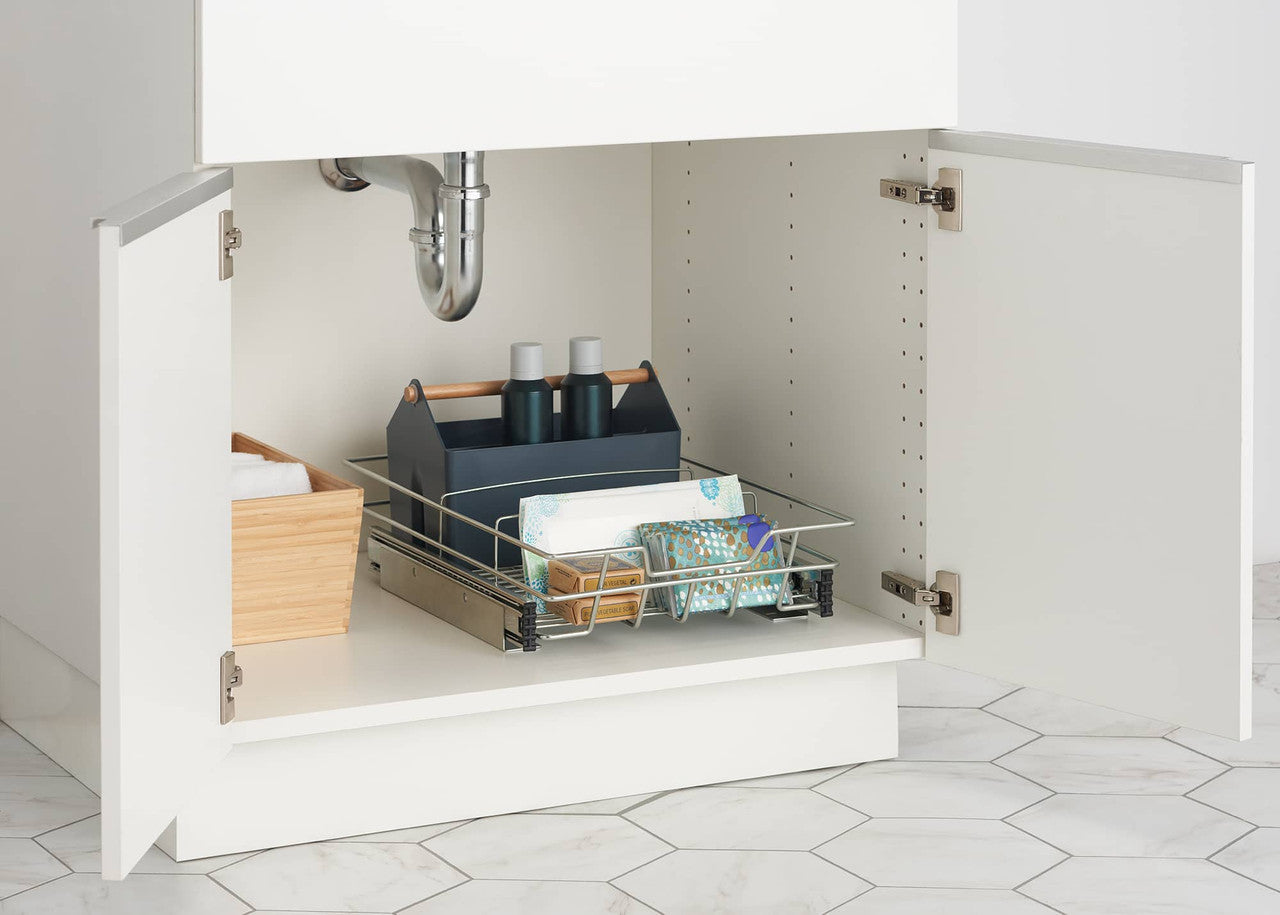 A chrome sliding organizer drawer is pulled partially out from under a white bathroom sink cabinet. The drawer holds various toiletries, including a blue caddy with bottles, a stack of white cloths, and a package of patterned washcloths.