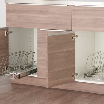 Two kitchen cabinets with light wood doors are partially open, revealing sliding metal organizers inside. The organizers have vertical dividers designed to hold bakeware or lids upright for easy access and storage.