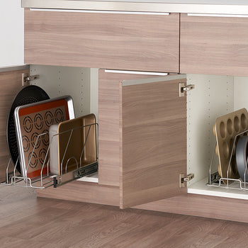 Two open kitchen cabinets with light brown wood doors reveal sliding metal organizers holding various baking pans and sheets. The organizers are mounted on rails allowing them to extend out for easy access to the stored items.