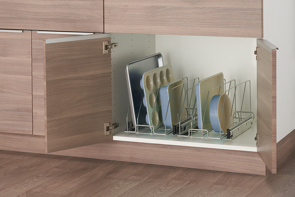 A low-angle, medium shot shows a partially opened light brown wooden kitchen cabinet revealing a chrome sliding bakeware organizer inside. The organizer holds several baking sheets, muffin tins, and round baking pans vertically, neatly arranged with dividers.