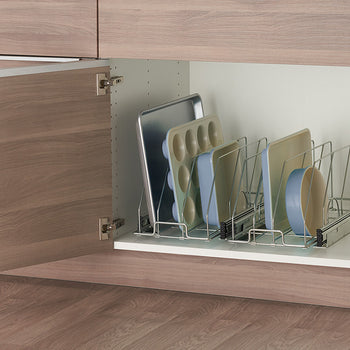 A low-angle, medium shot shows a partially opened light brown wooden kitchen cabinet revealing a chrome sliding bakeware organizer inside. The organizer holds several baking sheets, muffin tins, and round baking pans vertically, neatly arranged with dividers.
