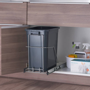 A dark gray rectangular trash can sits inside a light brown cabinet, mounted on a chrome sliding track that is partially extended. To the right of the trash can, also inside the cabinet, are cleaning supplies including spray bottles, a white bucket with sponges, and a box of trash bags.