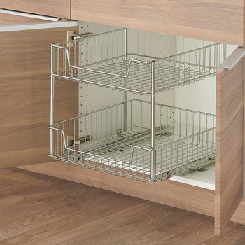 A close-up shows a two-tiered wire sliding drawer system installed within a light wood-toned cabinet. The drawers are empty, showcasing their chrome-like wire basket construction and the smooth sliding mechanism attached to the white interior of the cabinet.