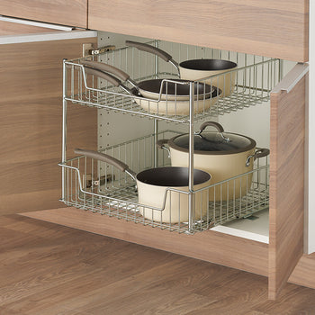 A two-tiered wire sliding drawer is partially extended from a light wood-grain cabinet. The drawer contains various beige pots and pans, neatly organized on each level.